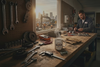 A man in a workbench area wearing a dark blue work jacket, working at a wooden table cluttered with tools, a steaming mug, and parts, with a city skyline visible through a window in the background.