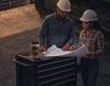 A man and a woman wearing white hard hats stand beside a dark tool chest, reviewing blueprints in a workshop or construction setting. A coffee mug sits on the chest.