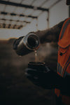 A person wearing an orange high-visibility safety vest and black gloves pours hot water from a stainless steel thermal mug into a black insulated cup, with steam rising in a warm, softly blurred outdoor setting.